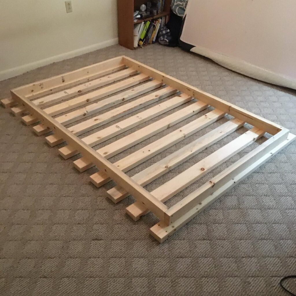 Wooden diy bed frame base with slats resting on a grey carpeted floor.