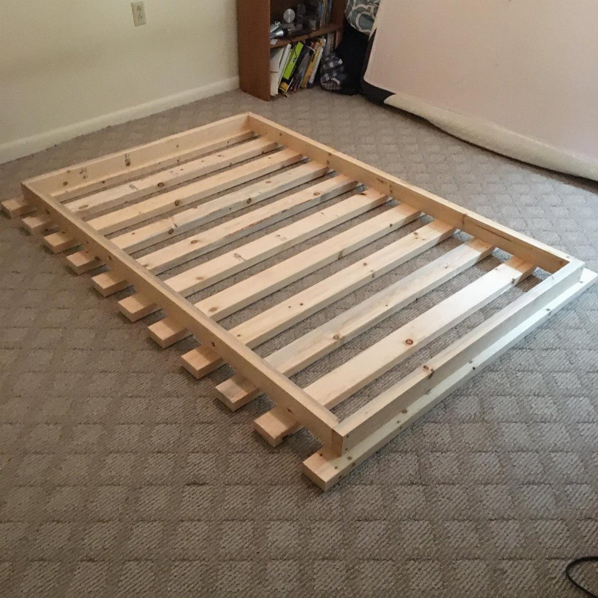 Wooden diy bed frame base with slats resting on a grey carpeted floor.