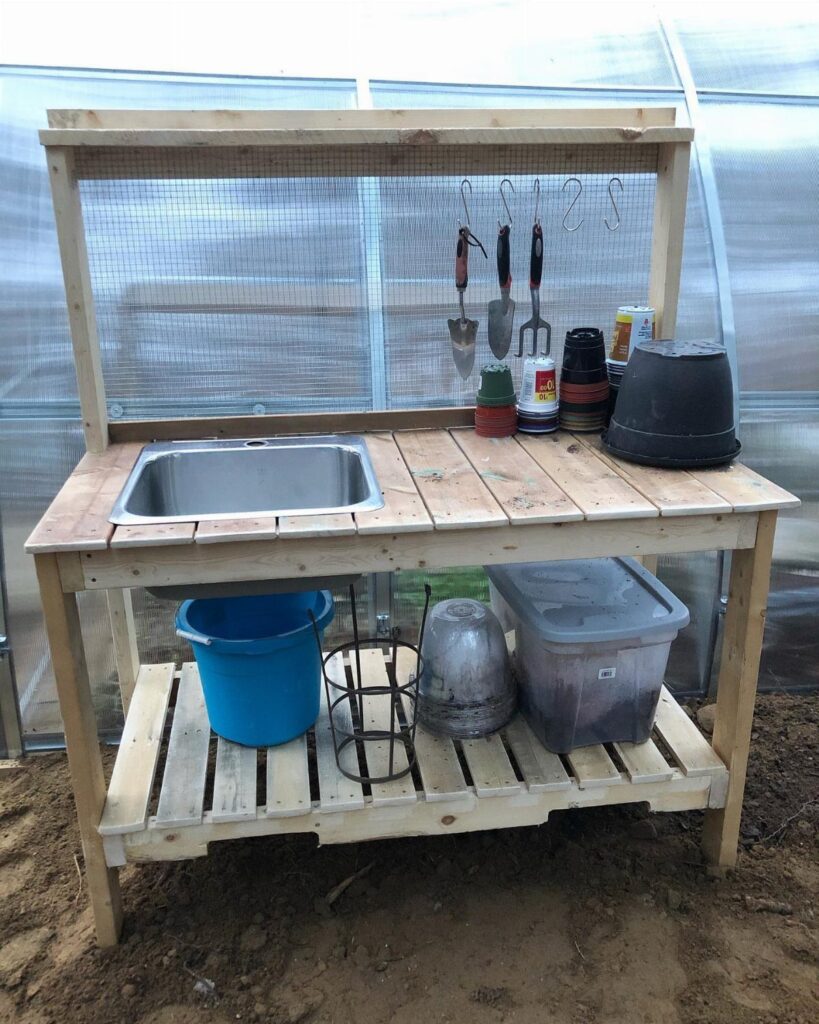Wooden potting bench with sink and tools inside a bright greenhouse setting.
