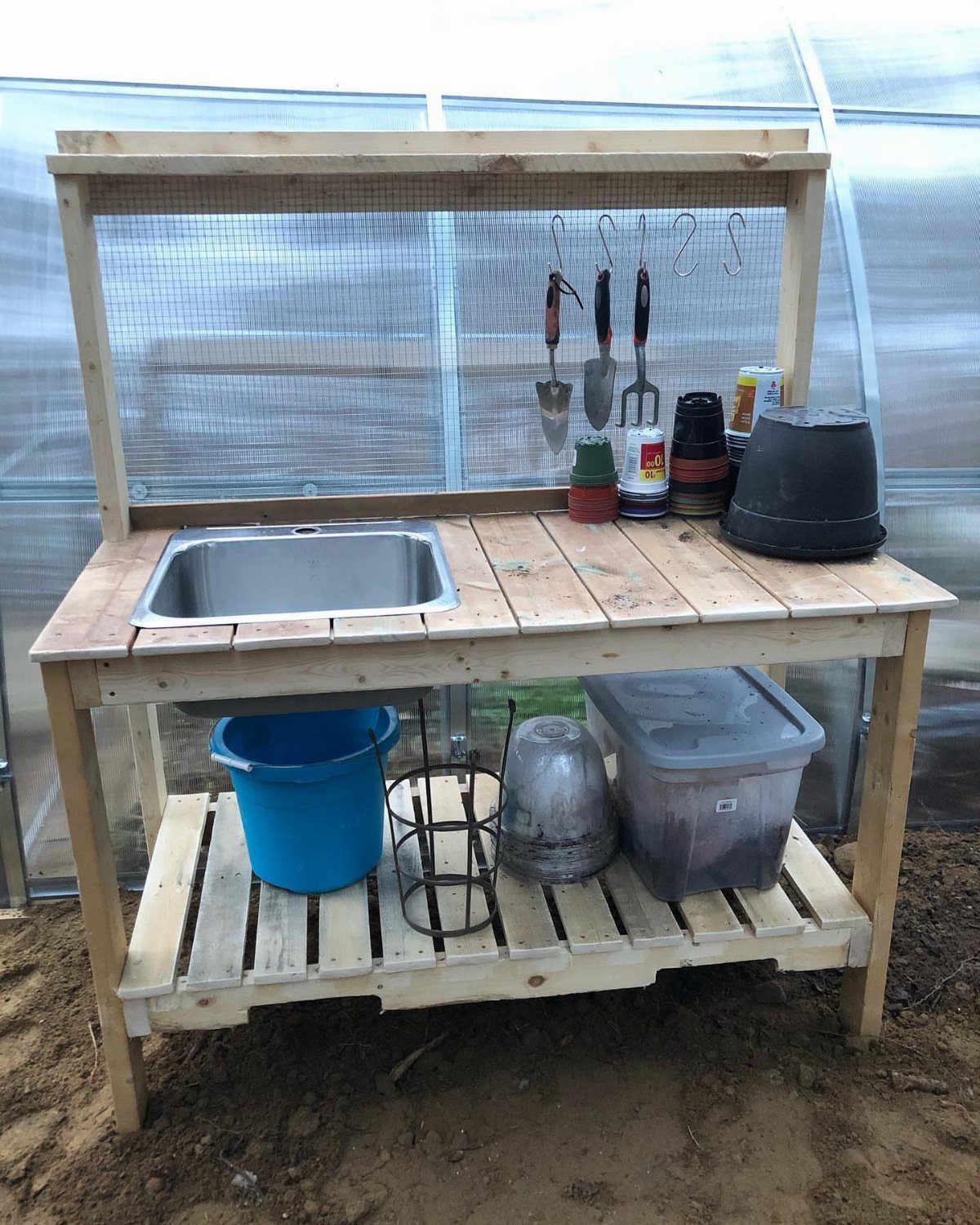 Wooden potting bench with sink and tools inside a bright greenhouse setting.