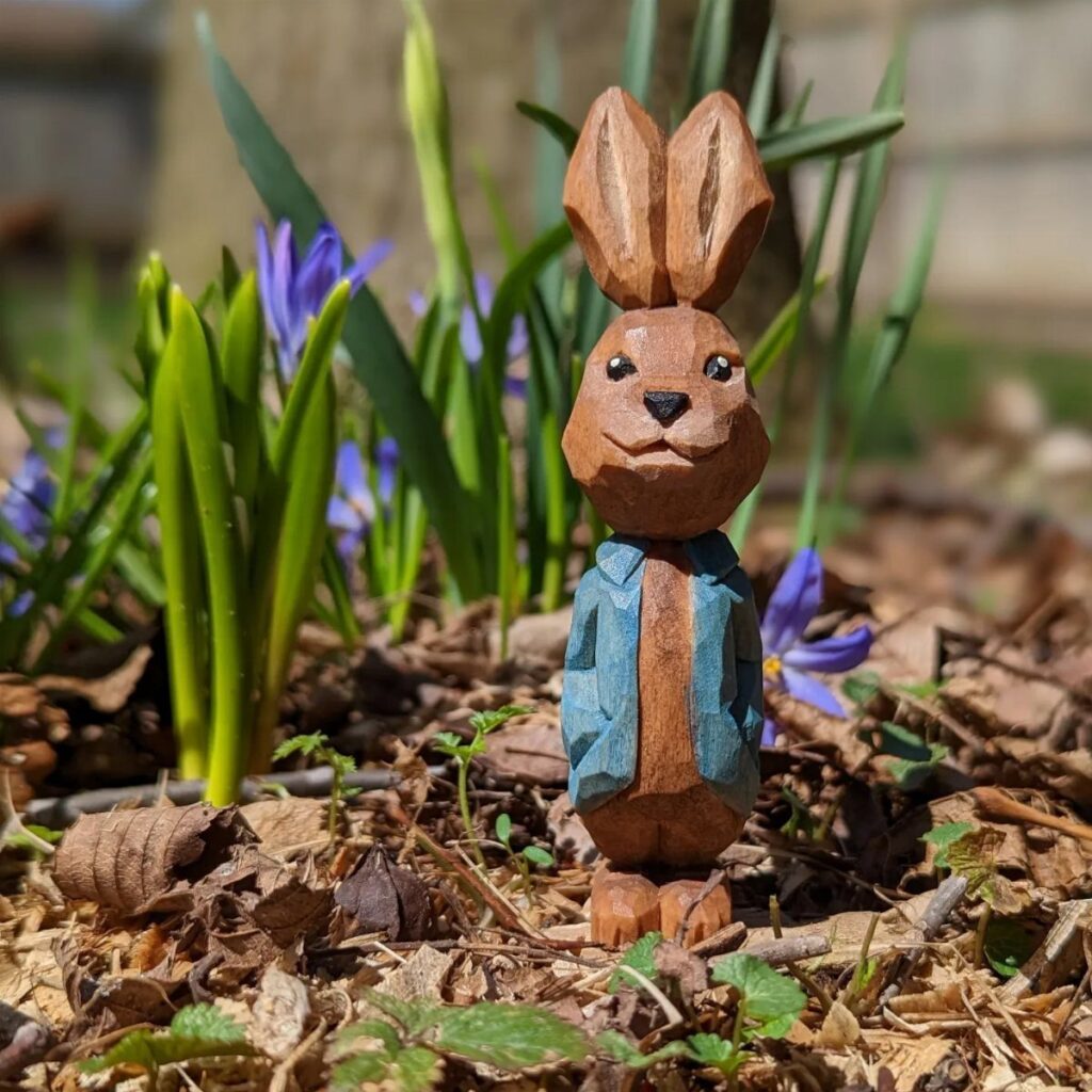 Hand-carved wooden rabbit wearing a blue jacket standing in a garden with purple flowers