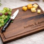 Personalized wooden cutting board with grapes, cheese, and a knife on marble.