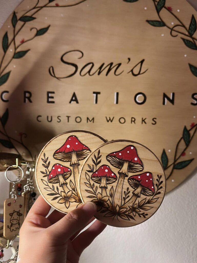 Two circular wooden coasters with wood-burned red mushroom designs held by a hand.