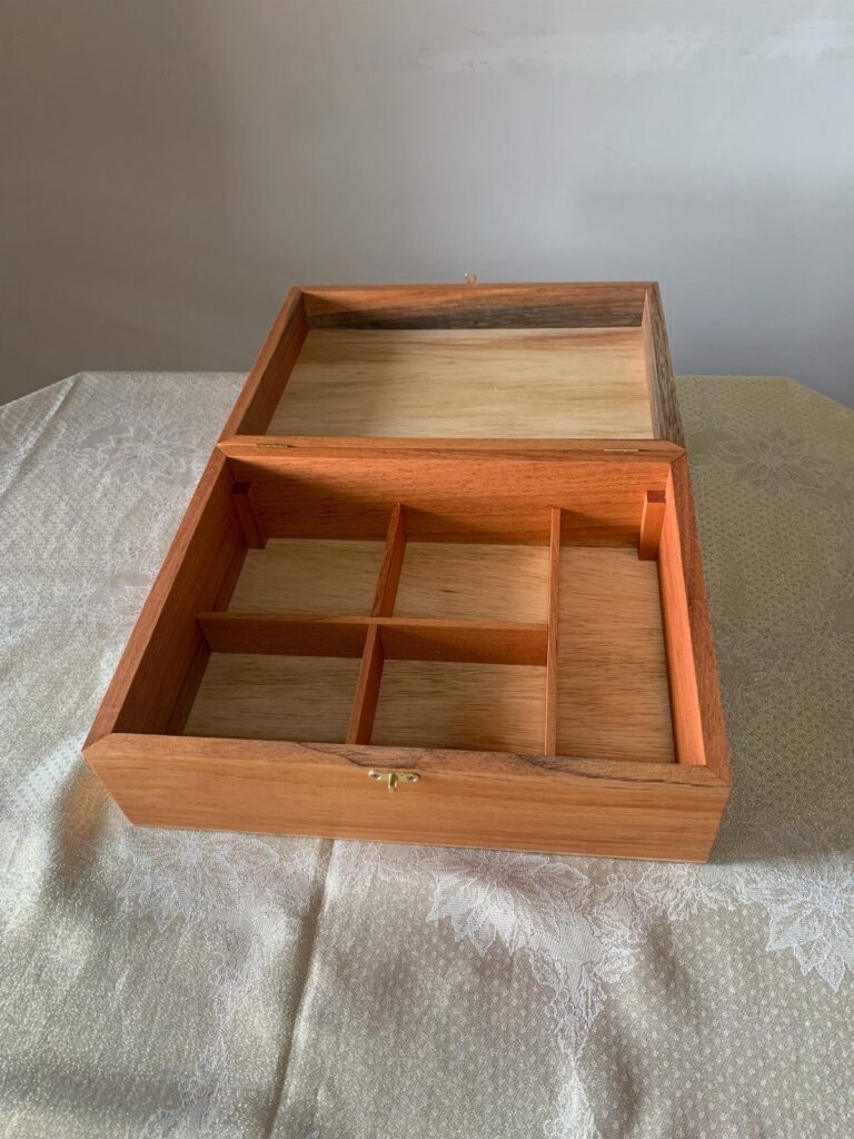 Open light brown wooden box with internal dividers on a silver fabric background.