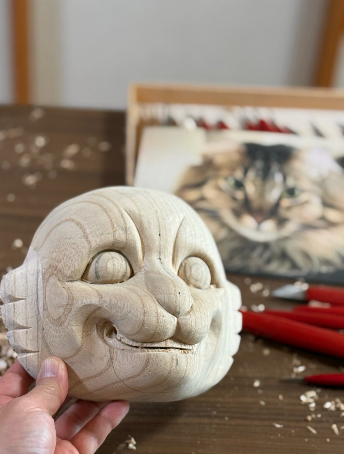 Carved wooden cat face held by a hand over a workbench with tools.