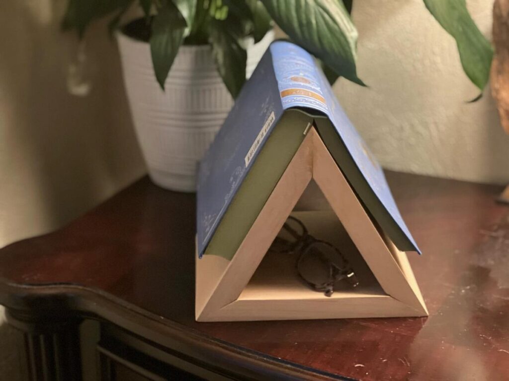 Wooden triangular book holder supporting an open blue book on a dark table.