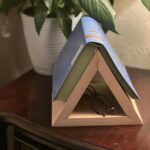 Wooden triangular book holder supporting an open blue book on a dark table.