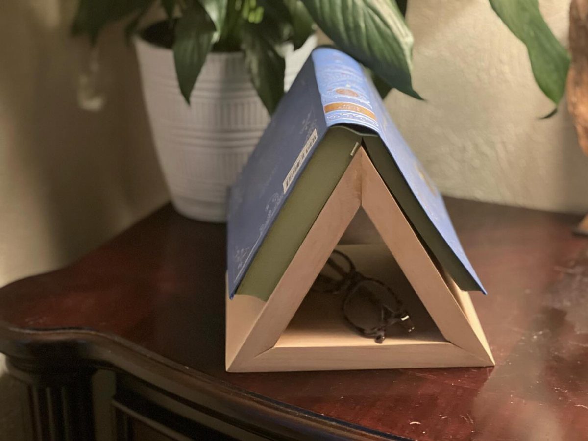 Wooden triangular book holder supporting an open blue book on a dark table.